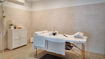 Een schone, moderne spa-ruimte met een massagetafel, witte handdoeken en een kastje met decoratieve objecten.