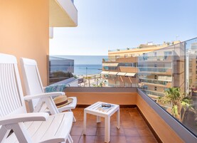 Een balkon met witte stoelen en een kleintje tafeltje, met uitzicht op een strand en andere gebouwen.