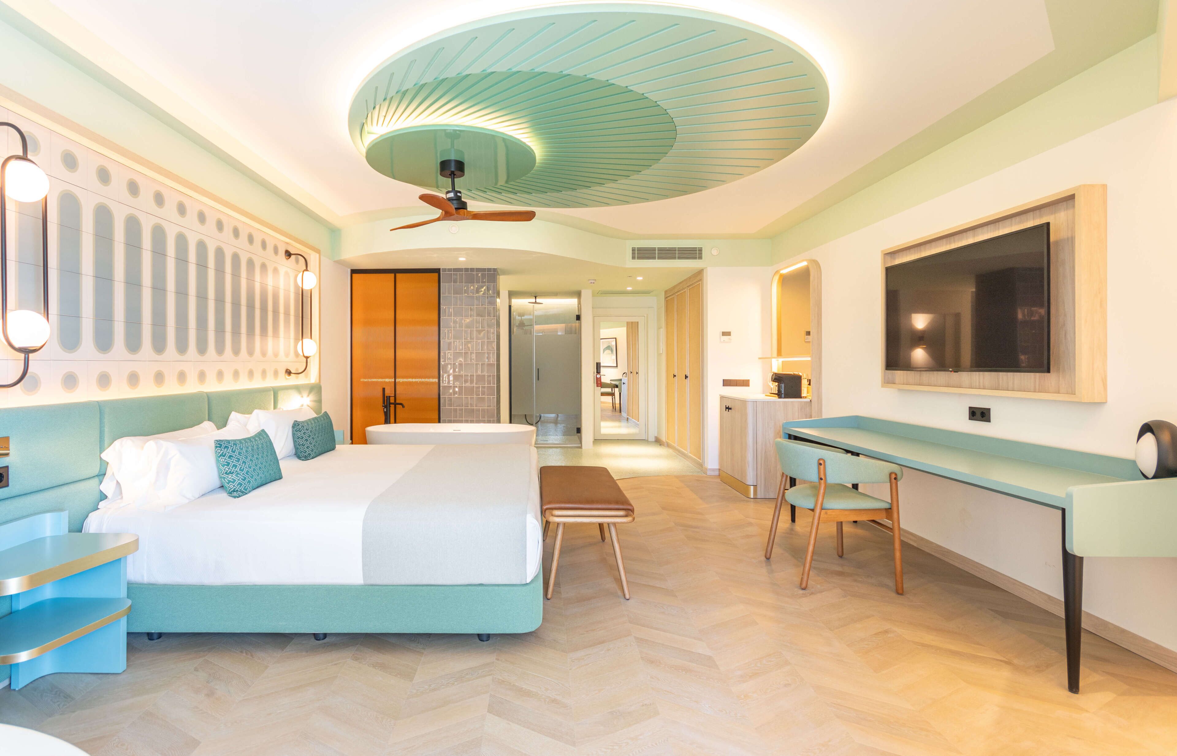 A modern hotel room with a large bed, a desk, a chair, a television, and a ceiling fan.