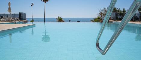 Piscina azul y transparente con vista al océano, una palmera y sillas de descanso.