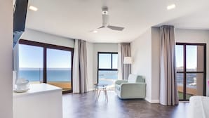 Una habitación de hotel moderna con un ventanal grande con vistas al mar, un ventilador de techo, una zona de estar con una mesa y un armario blanco con una taza.