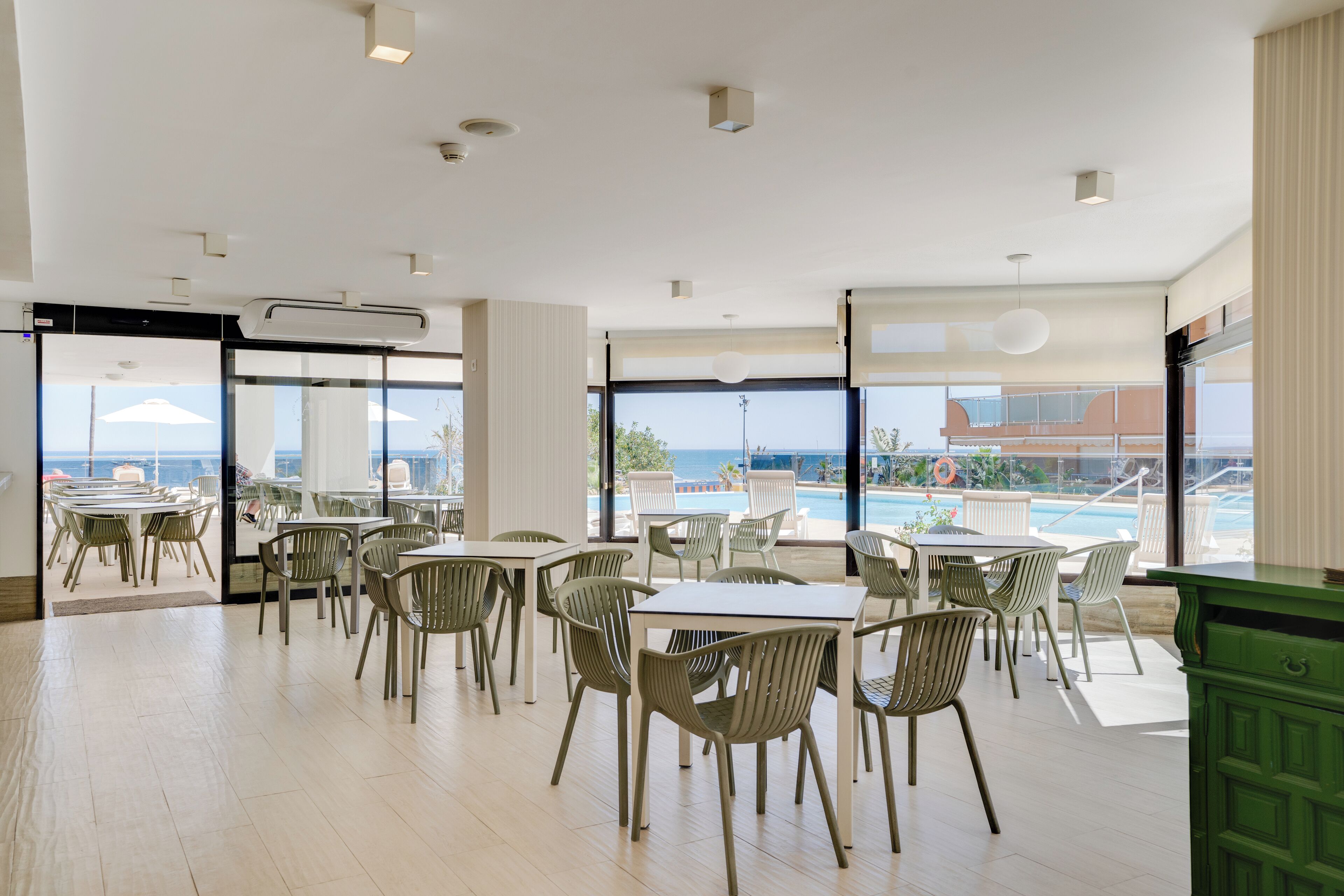 A spacious indoor dining area with tables and chairs, large windows offering a view of the pool and ocean, and an air conditioning unit on the ceiling.