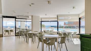 A spacious indoor dining area with tables and chairs, large windows offering a view of the pool and ocean, and an air conditioning unit on the ceiling.