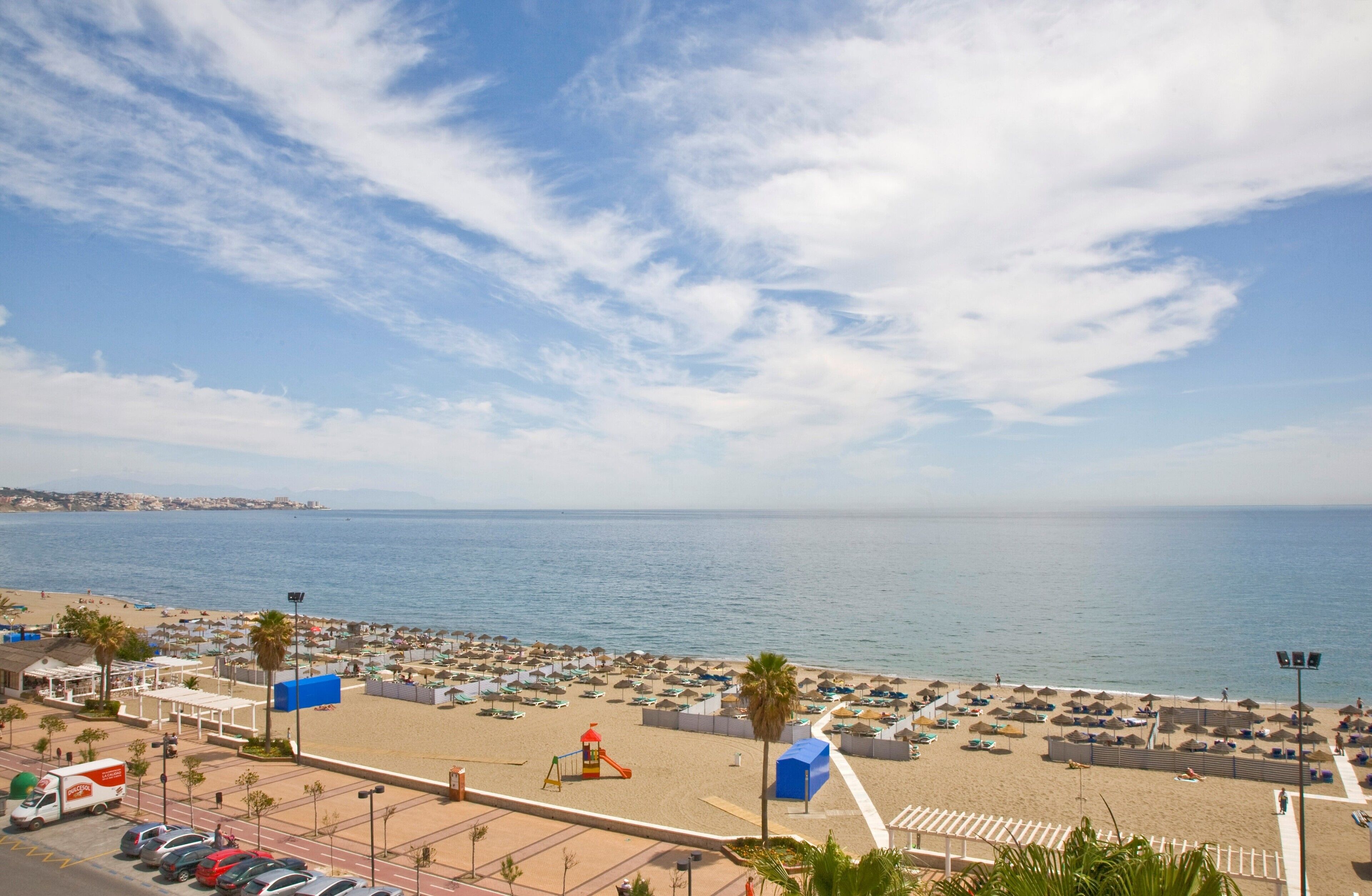 A beach with umbrellas and sun loungers, a playground, and a parking area with cars.