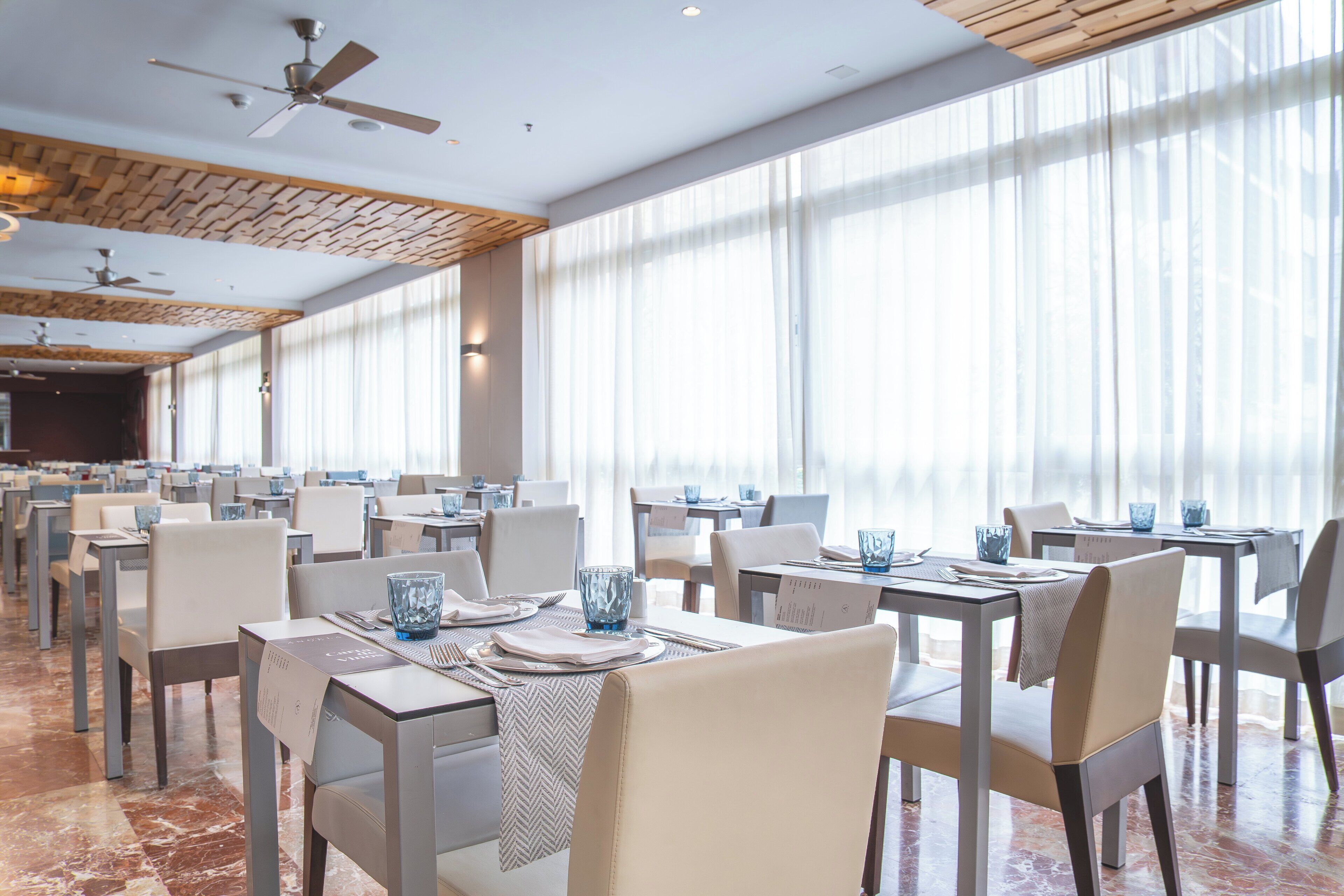 Un'ampia sala da pranzo con diversi tavoli e sedie, grandi finestre con tende leggere e ventilatori a soffitto.