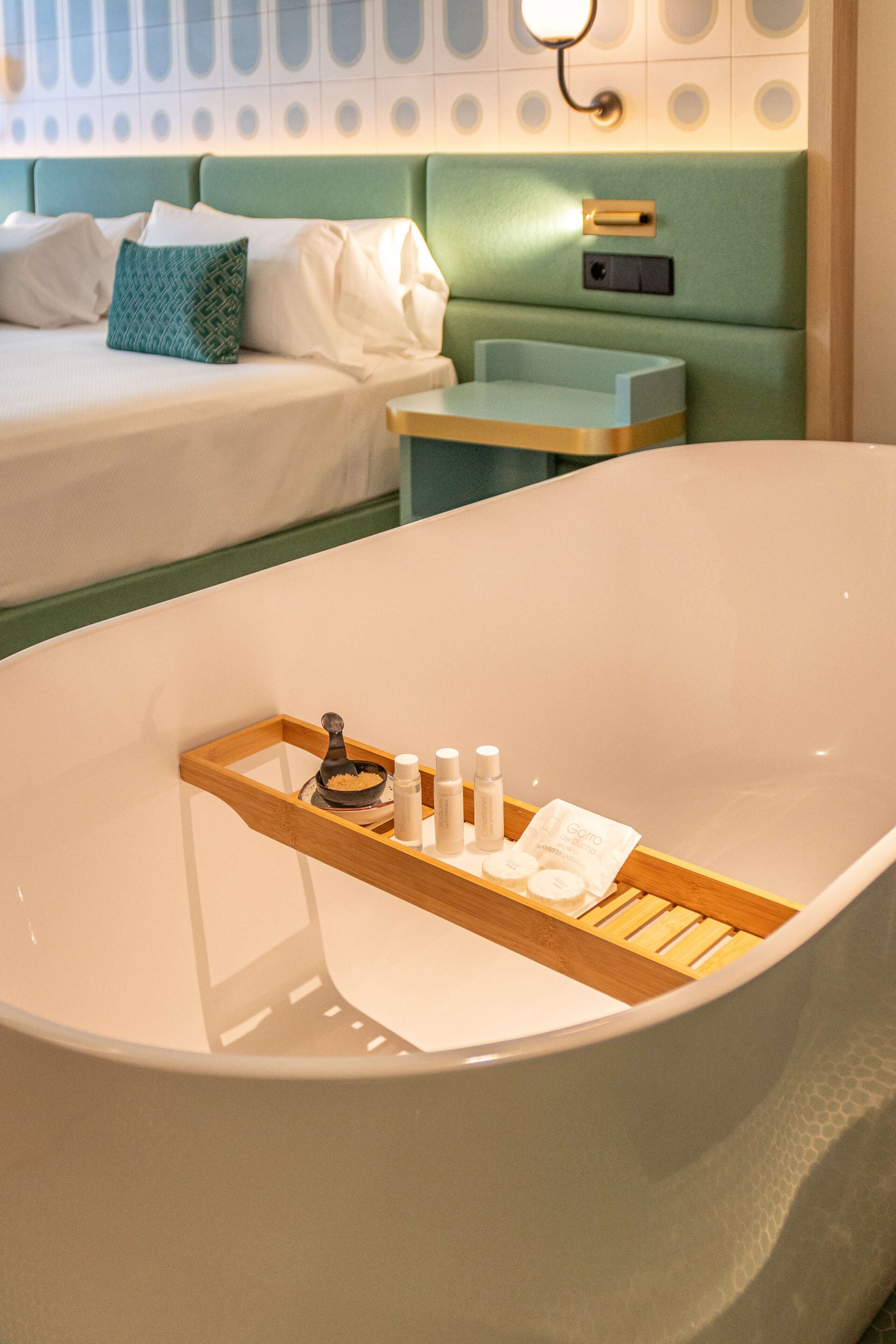 A modern bathroom with a freestanding bathtub, a wooden tray with toiletries, and a bed with a green headboard in the background.