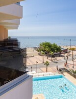 Zona de piscina con agua azul cristalina, vista a la playa, y una calle con palmeras y edificios.