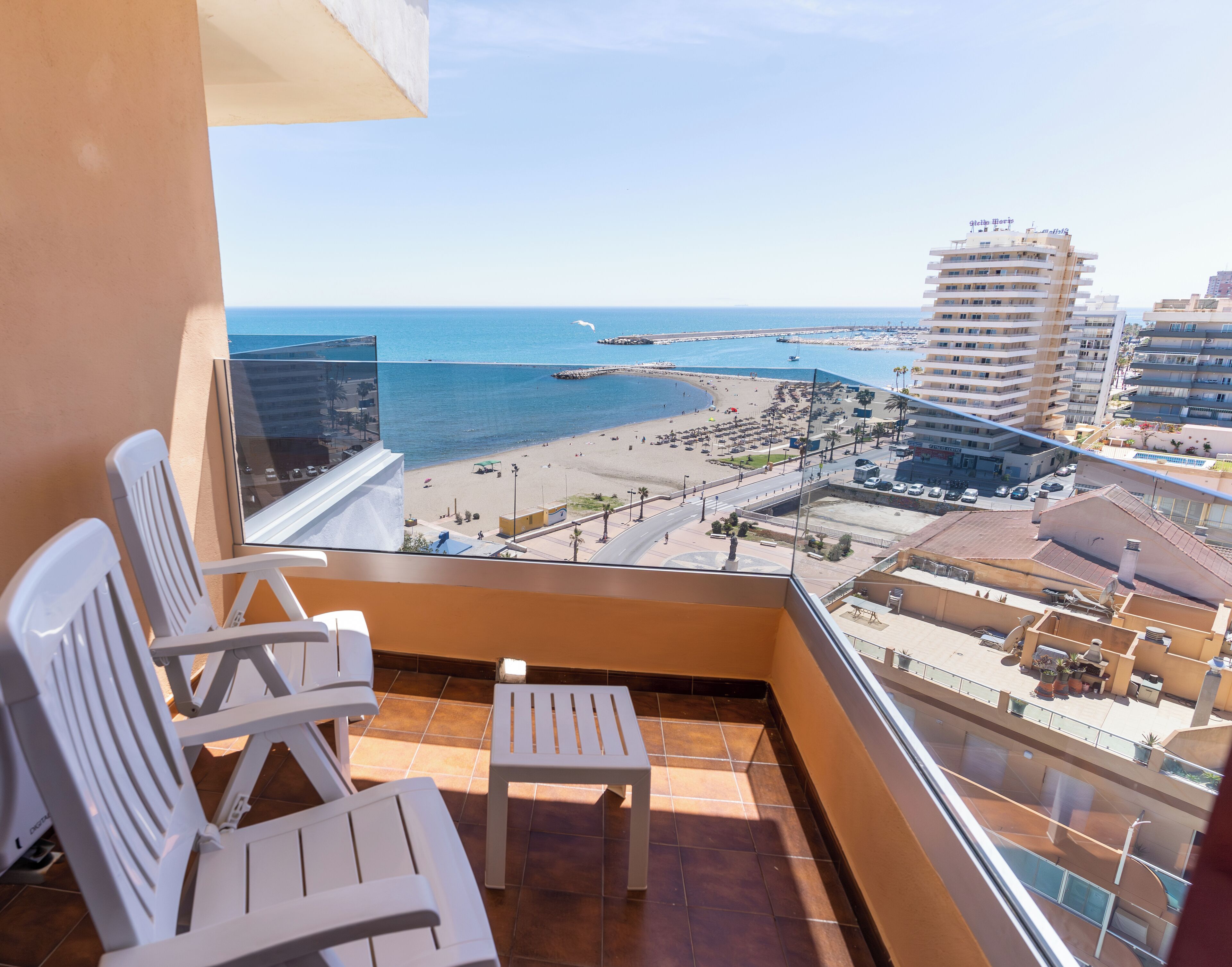 A balcony with white chairs and a small table, overlooking a beach and a cityscape with buildings and a pier.