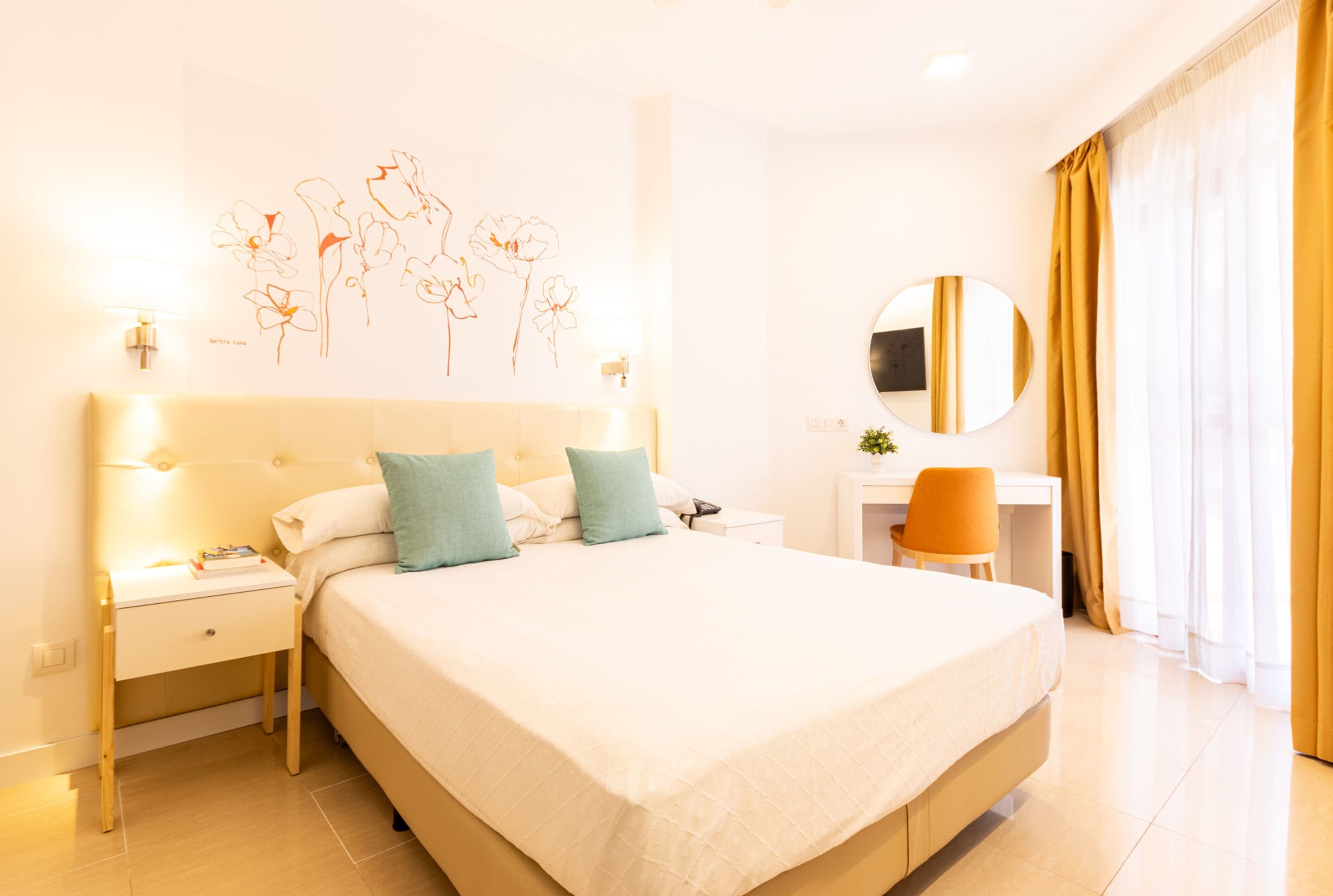 A hotel room with a large bed, bedside tables, a desk, a chair, a round mirror, and a wall art piece.
