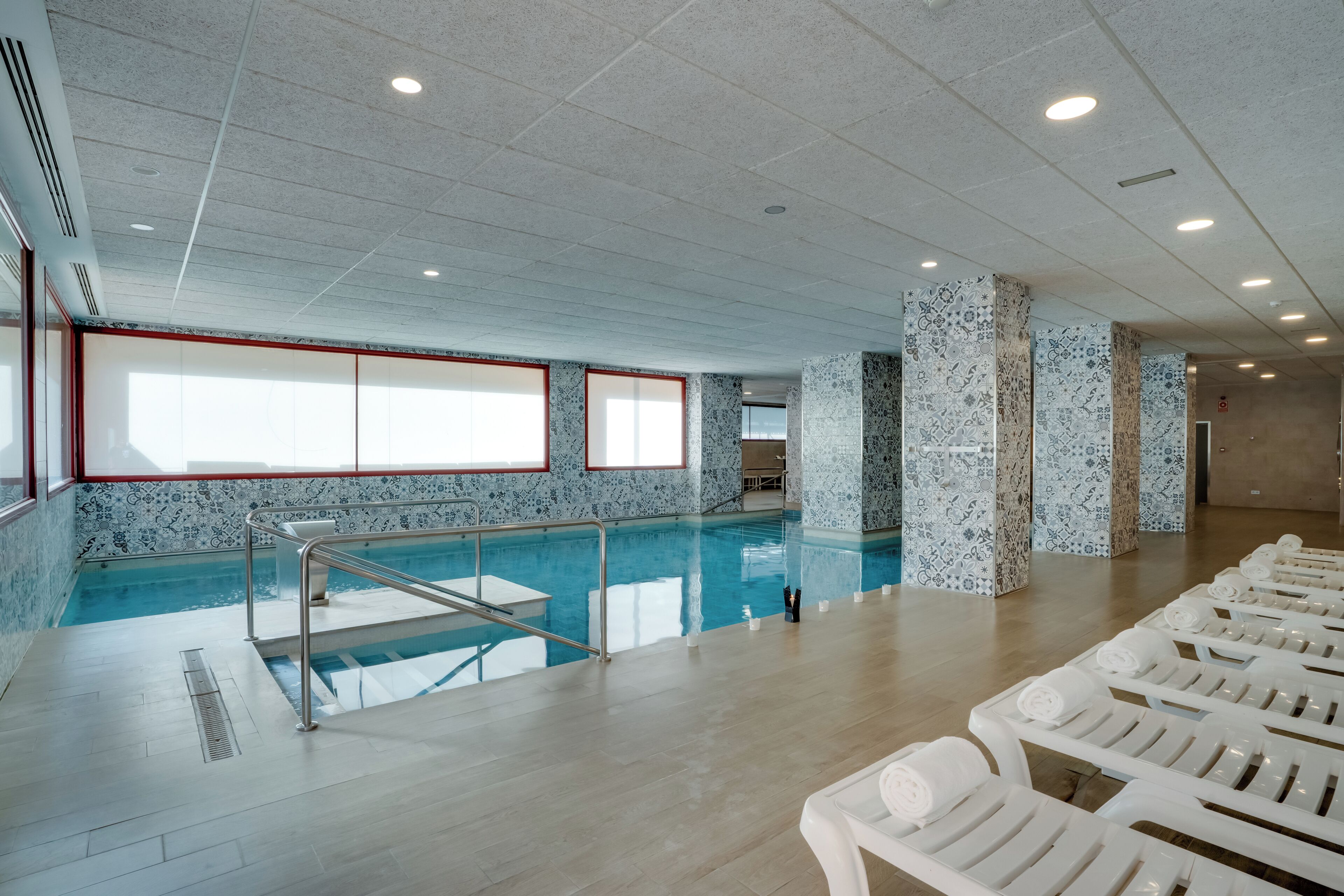Una piscina con pared de azulejos y una ventana. Hay sillas de descanso y suelo de baldosas.