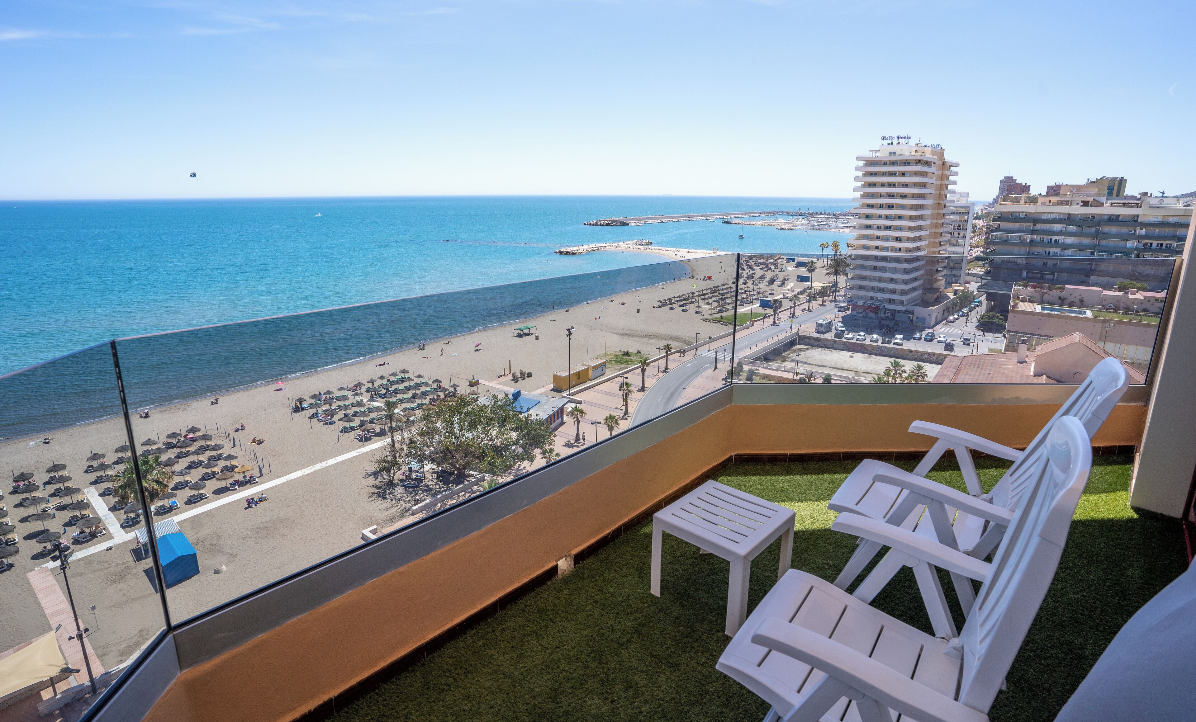 A balcony with a view of a beach and ocean. There are white chairs and a small table.