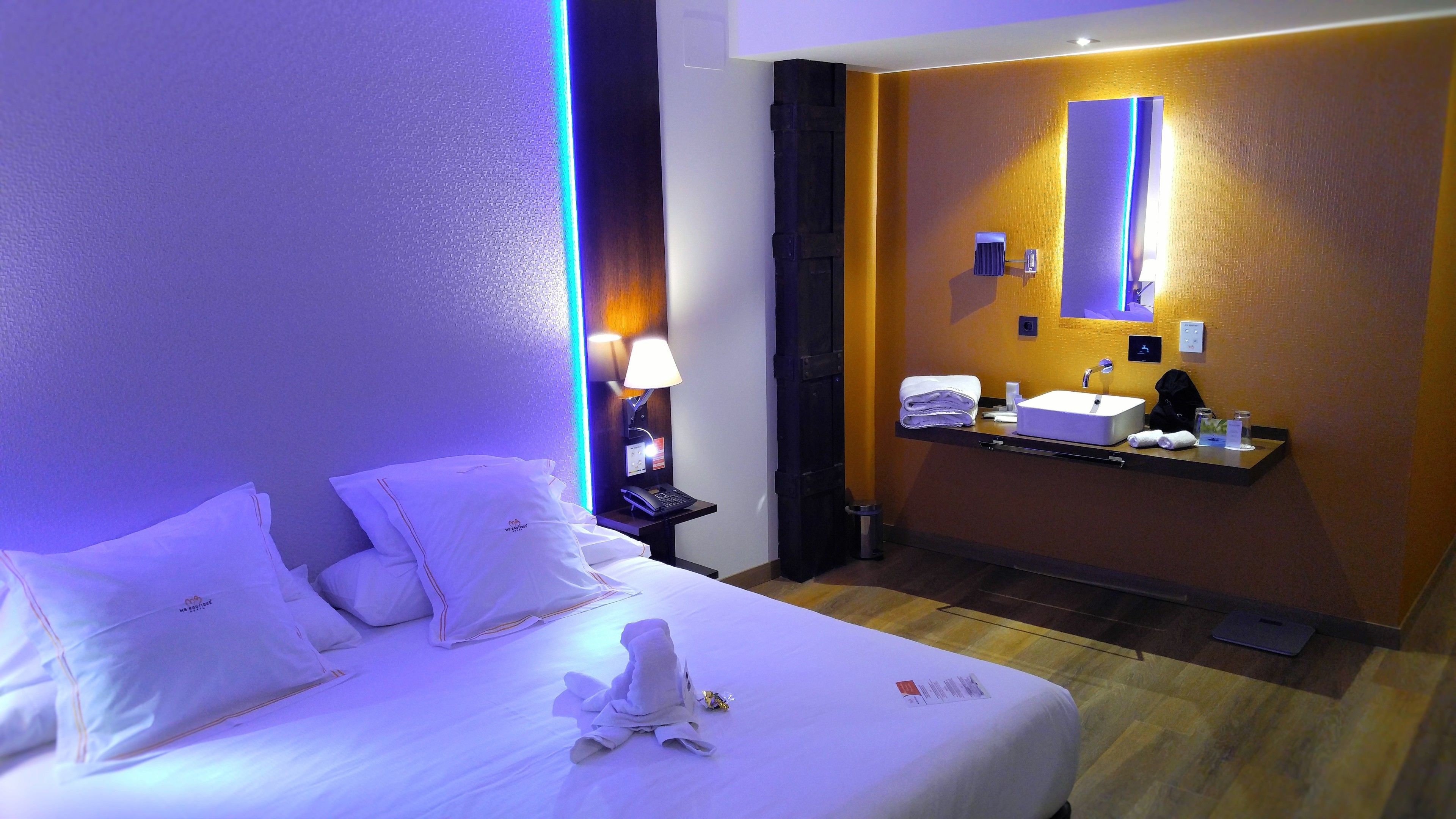 A hotel room with a bed, a bedside table with a lamp, a mirror, a sink, and a towel on the bed.