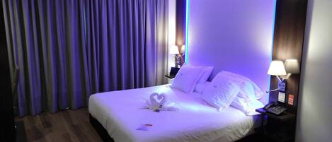 Habitación de hotel con una cama bien hecha, una computadora portátil sobre la mesita de noche y una toalla con forma de corazón en la cama.
