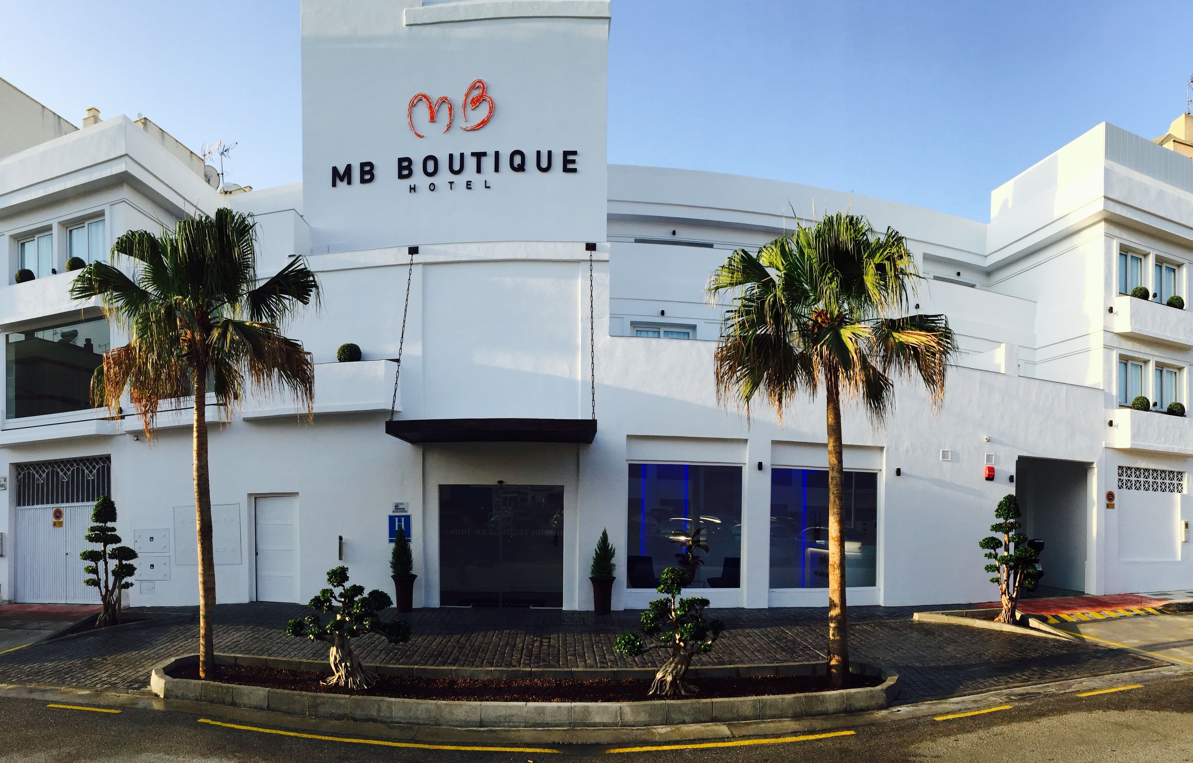 Ein modernes MB Boutique Hotel mit einem gut sichtbaren MB-Logo, heller Fassade und Palmen im Vordergrund.