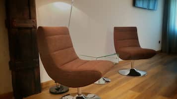 Two brown upholstered chairs with metal bases, a glass table, and a black floor lamp in a room with wooden flooring and a white wall.