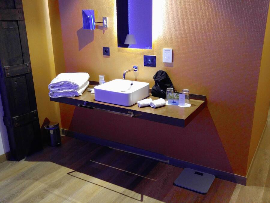 A hotel bathroom with a sink, mirror, towel dispenser, and a scale on the floor.