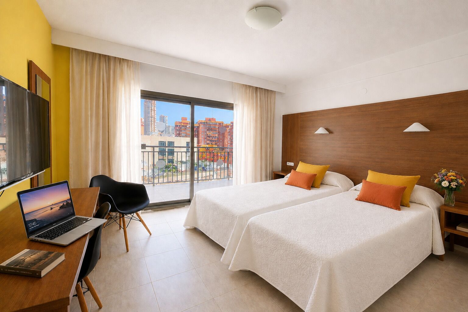 A hotel room with two beds, a desk with a laptop, a chair, a TV, and a balcony with a city view.