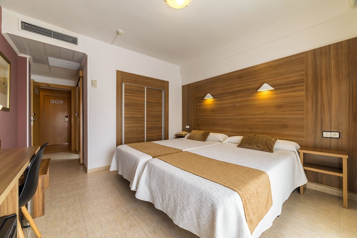 Ein Hotelzimmer mit zwei Betten, einem Holzkopfstück, einem Nachttisch, einem Schreibtisch mit Stuhl und einer Tür zu einem weiteren Raum.