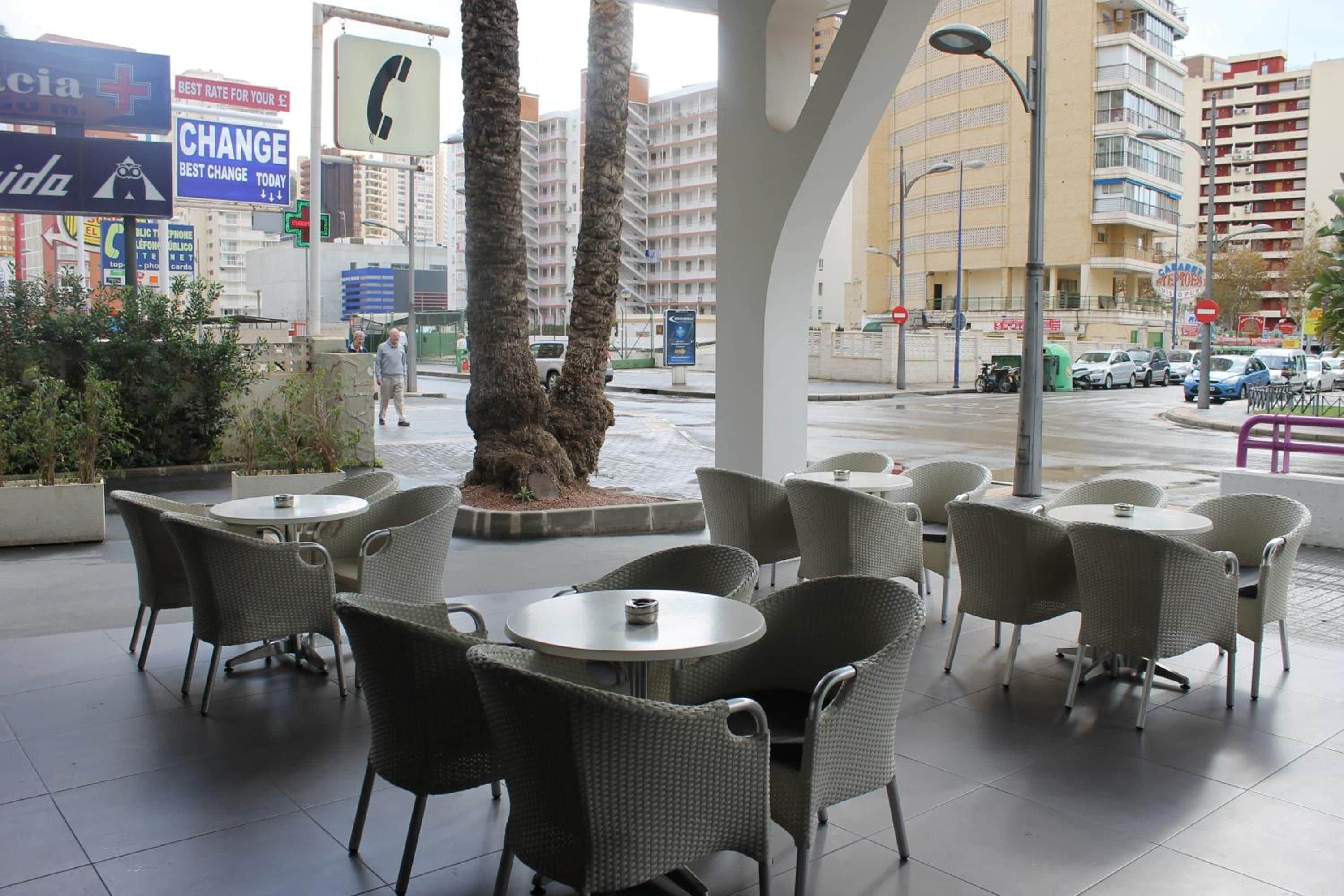 An outdoor seating area with wicker chairs and tables, palm trees, and a view of a city street with buildings and cars.