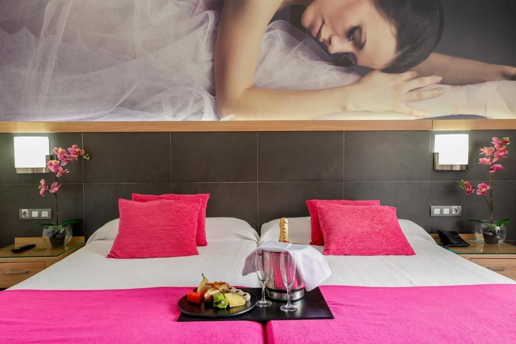 A hotel room with a large bed, two pink pillows, a champagne bottle, two glasses, and a fruit plate on a black tray.