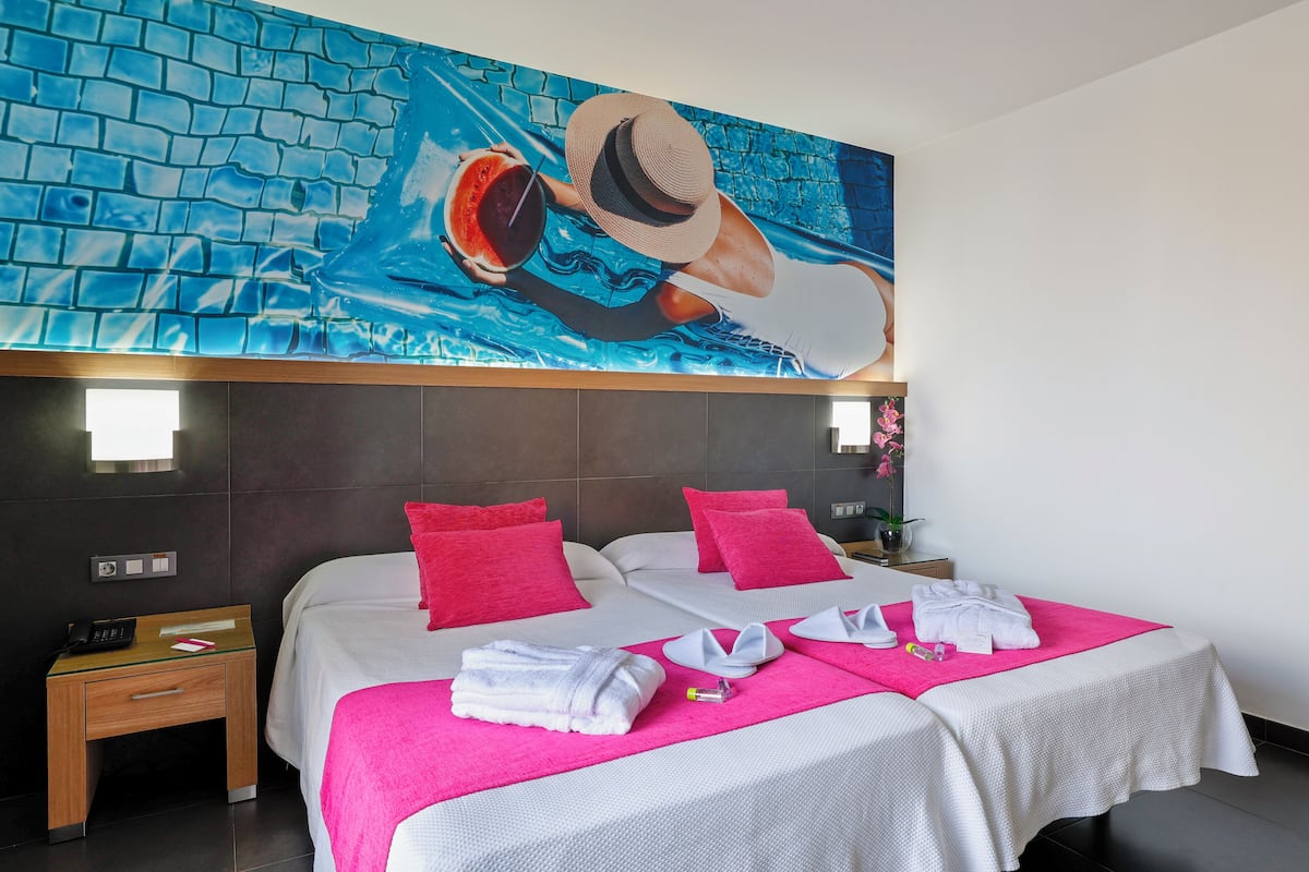 A hotel room with two beds, each with a pink pillow, a glass nightstand with a phone, and a bright pink towel.