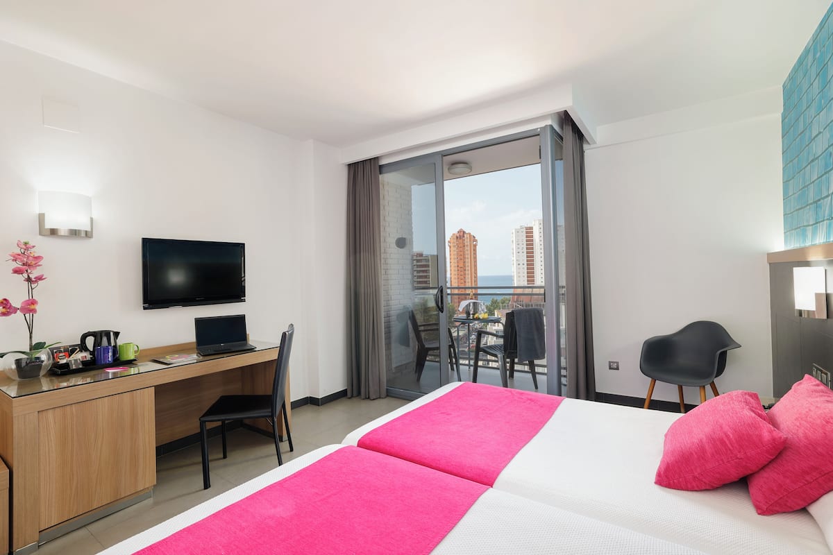 A hotel room with a bed, a desk with a laptop, a TV, and a balcony with a view of buildings.