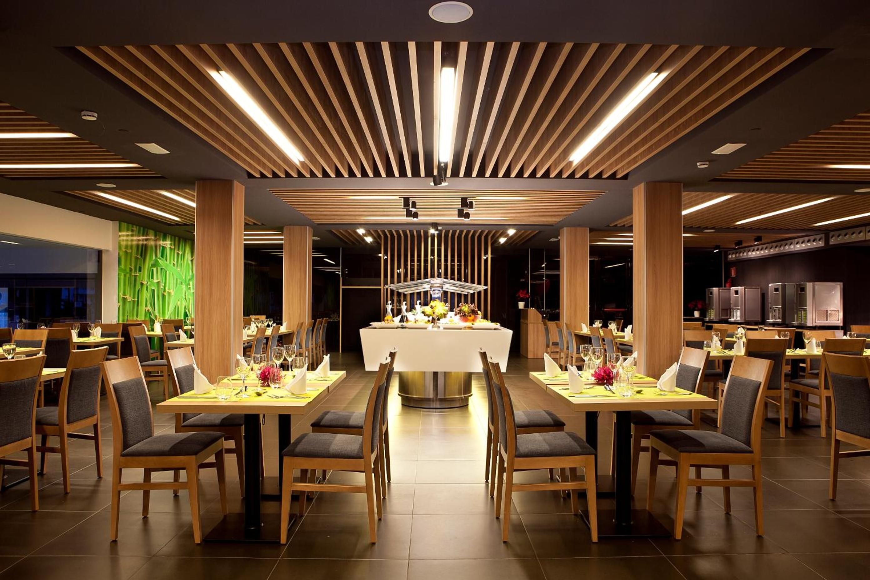 A modern restaurant with a central dining area, wooden panel ceiling, and multiple tables with chairs.