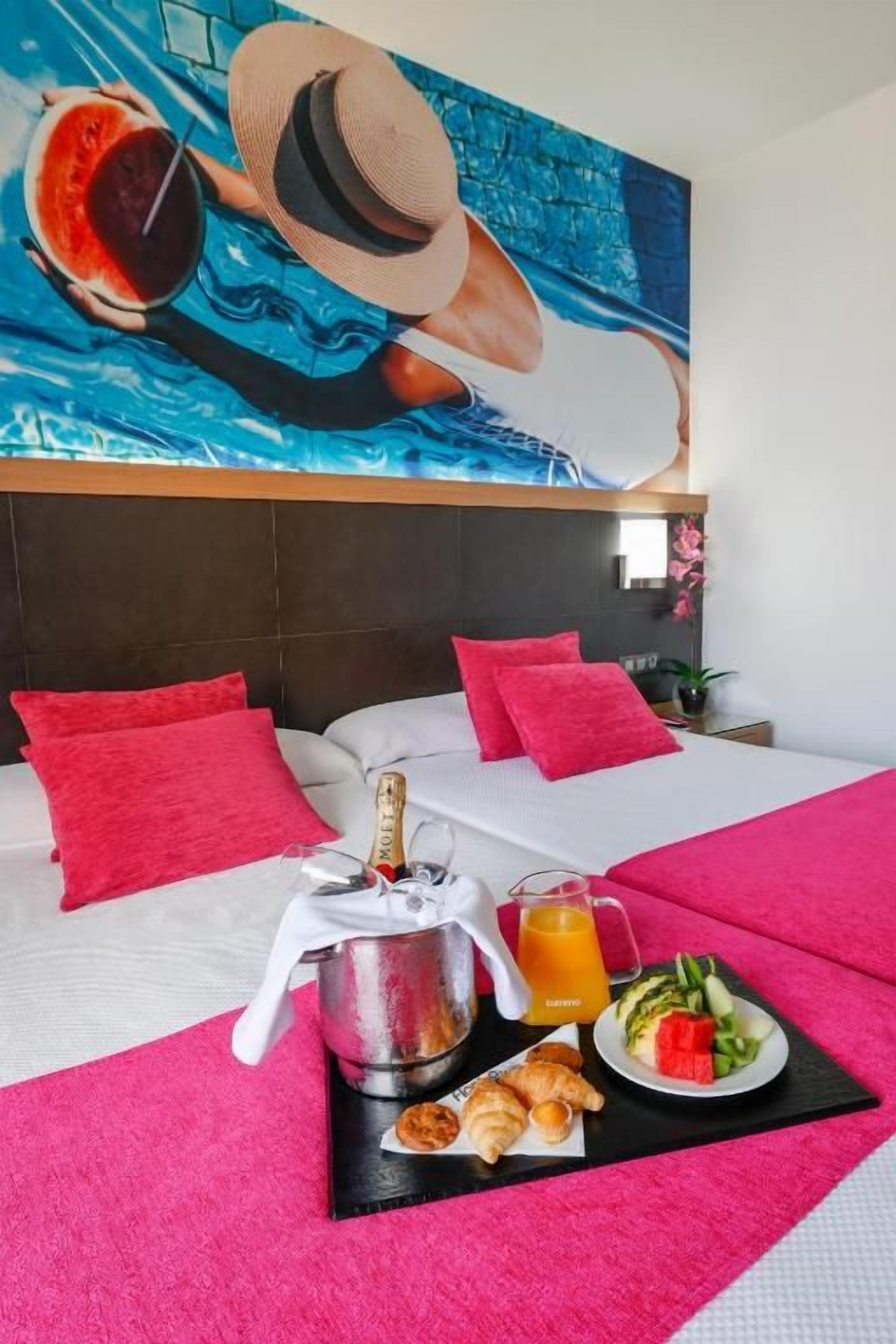 A hotel room with a bed, a breakfast tray, a bottle of champagne, and a painting of a person in a hat.