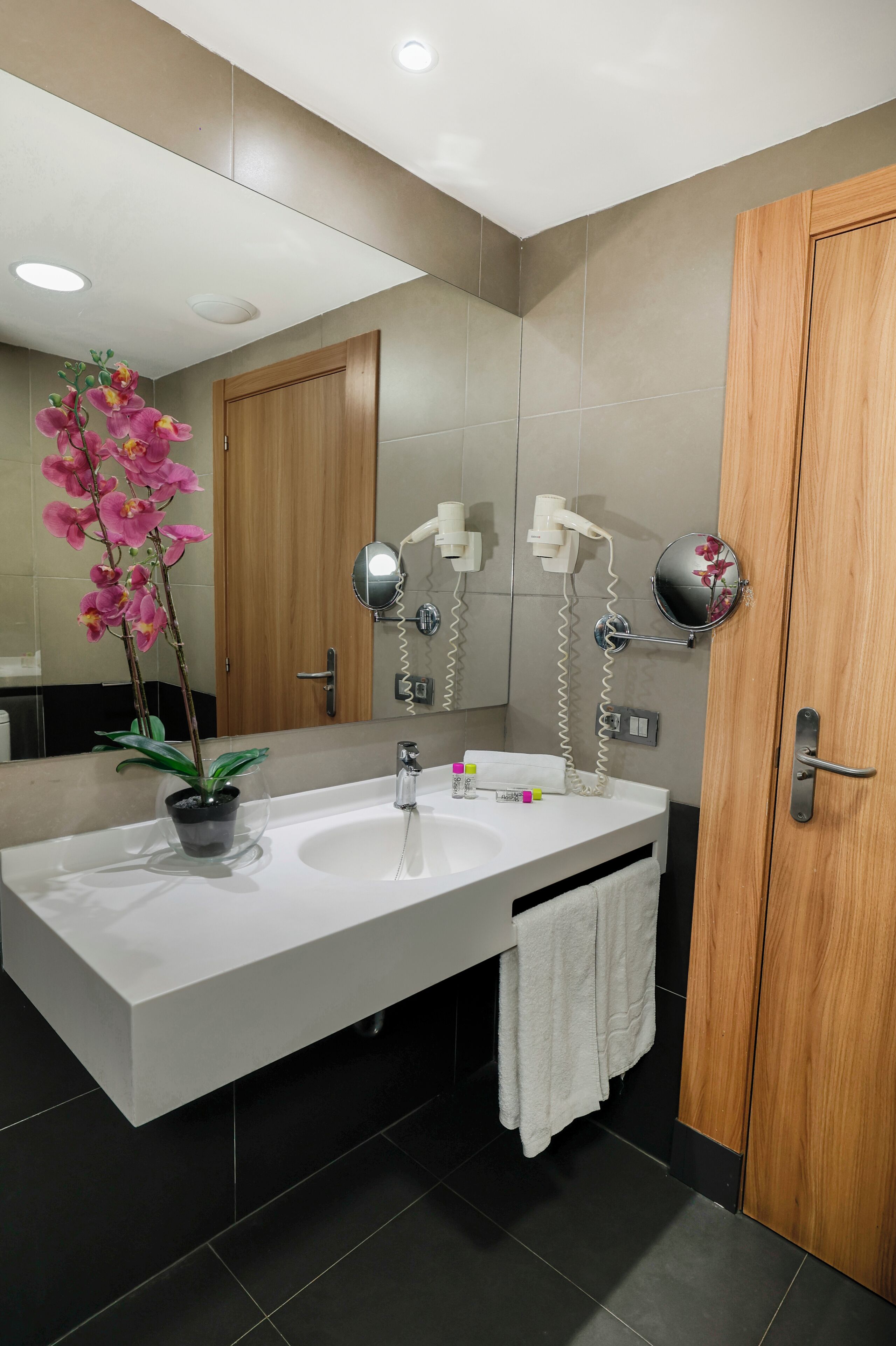 A modern bathroom with a white sink, two mirrors, a potted orchid, and a wooden door.