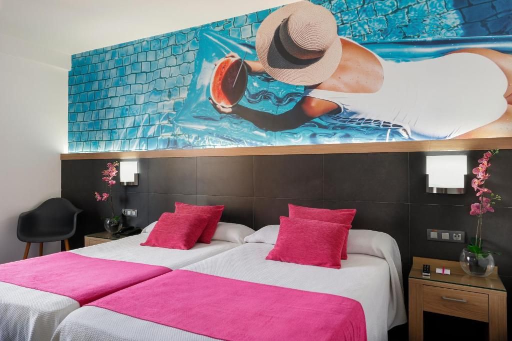 Habitación de hotel con un gran mural de una piscina, una cama con ropa de cama rosa y una mesita de noche con un florero.
