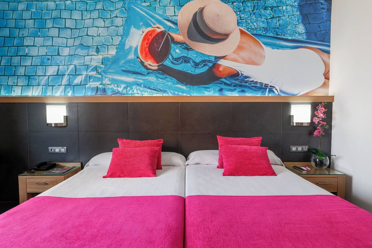 A hotel room with two beds, each with a red pillow and a white bedspread. There is a large mural of a person swimming on the wall above the beds.