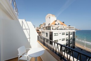 Un balcon avec une chaise et une table blanches, donnant sur une plage et des immeubles.