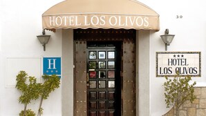 De ingang van Hotel Los Olivos, met een beige luifel, een dubbele deur en een balkon erboven.