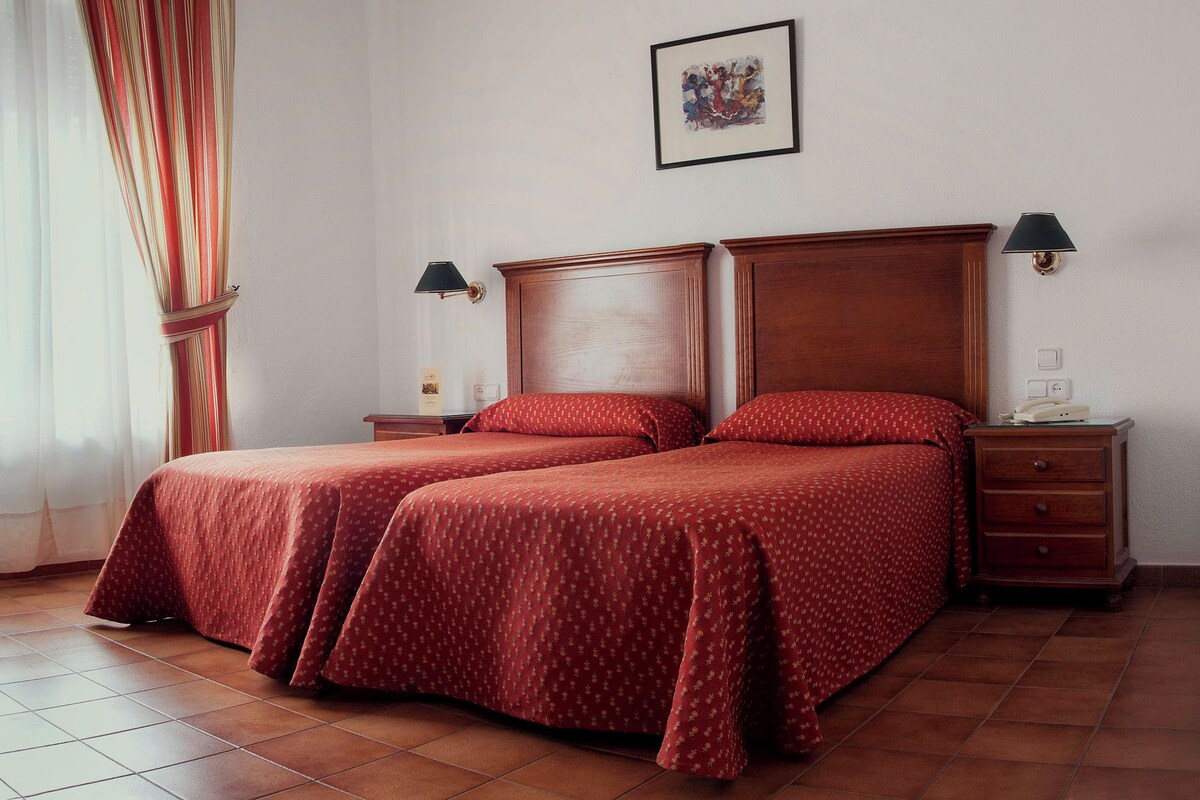 Habitación de hotel con dos camas, cabecera de madera, mesita de noche con lámpara y un cuadro en la pared.
