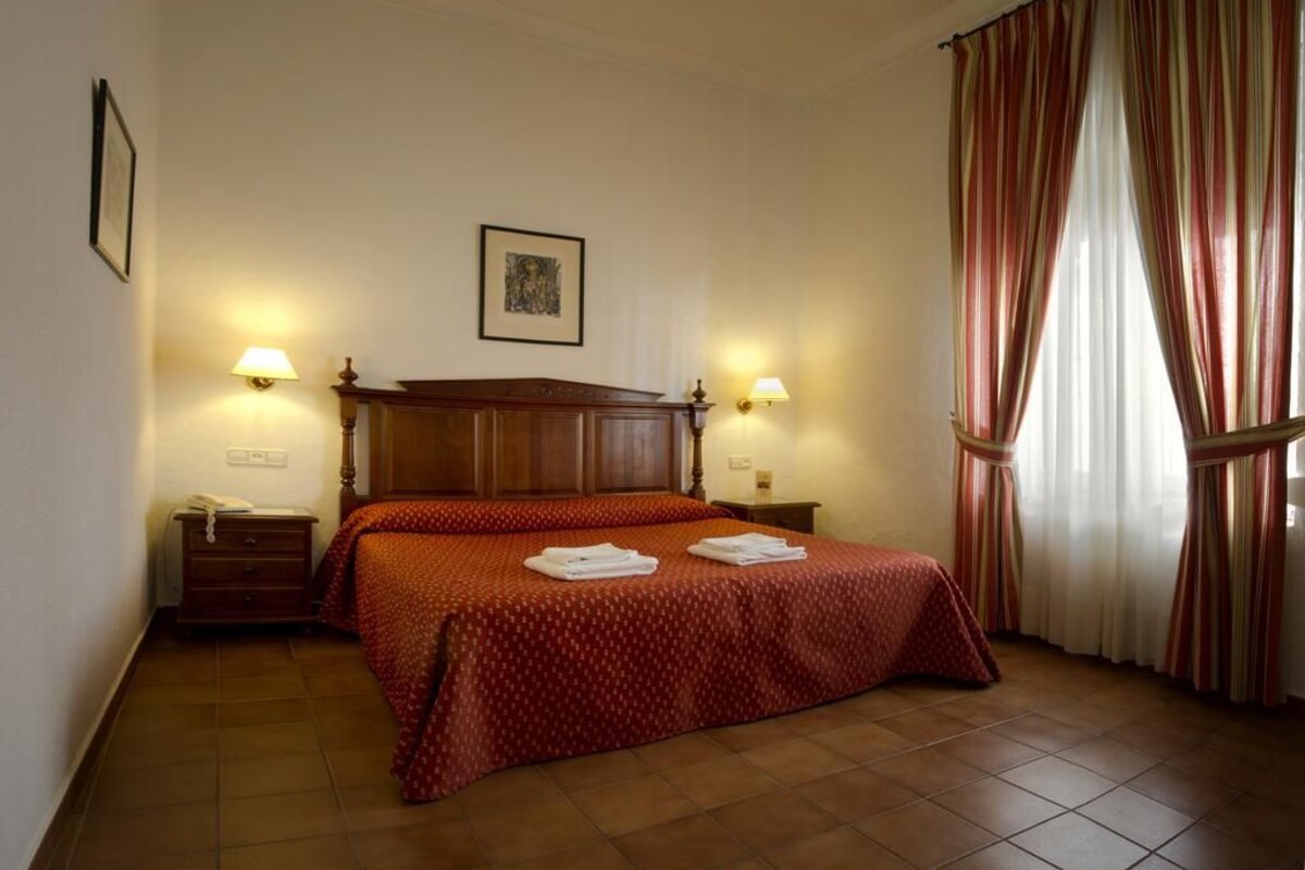 Una habitación de hotel con una cama doble, dos lámparas de noche, un cabecero de madera, una mesita de noche pequeña con una lámpara, y un ventanal con cortinas.