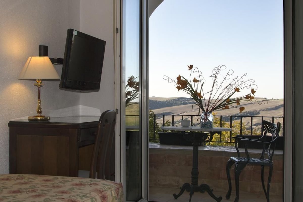 Una habitación con escritorio, lámpara, televisión montada en la pared, silla, mesita con un jarrón de flores y un balcón con vistas a las colinas.