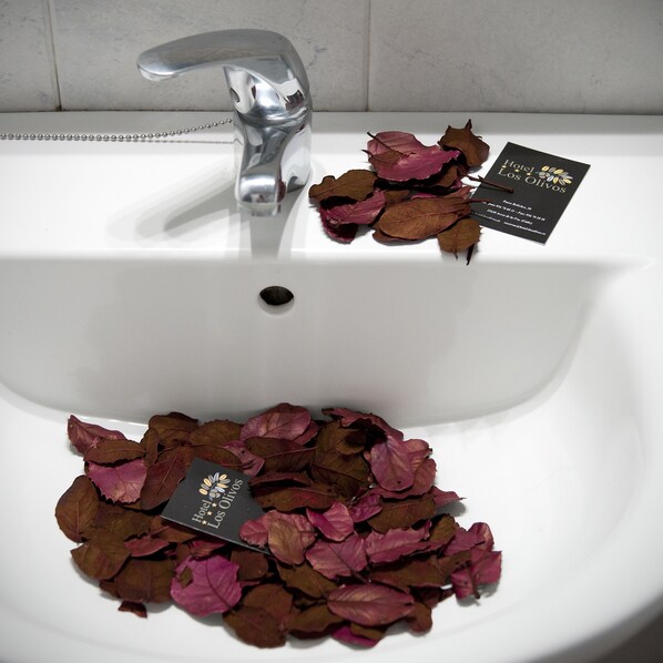 A white sink with a silver faucet, surrounded by dried rose petals and two black business cards with gold text.