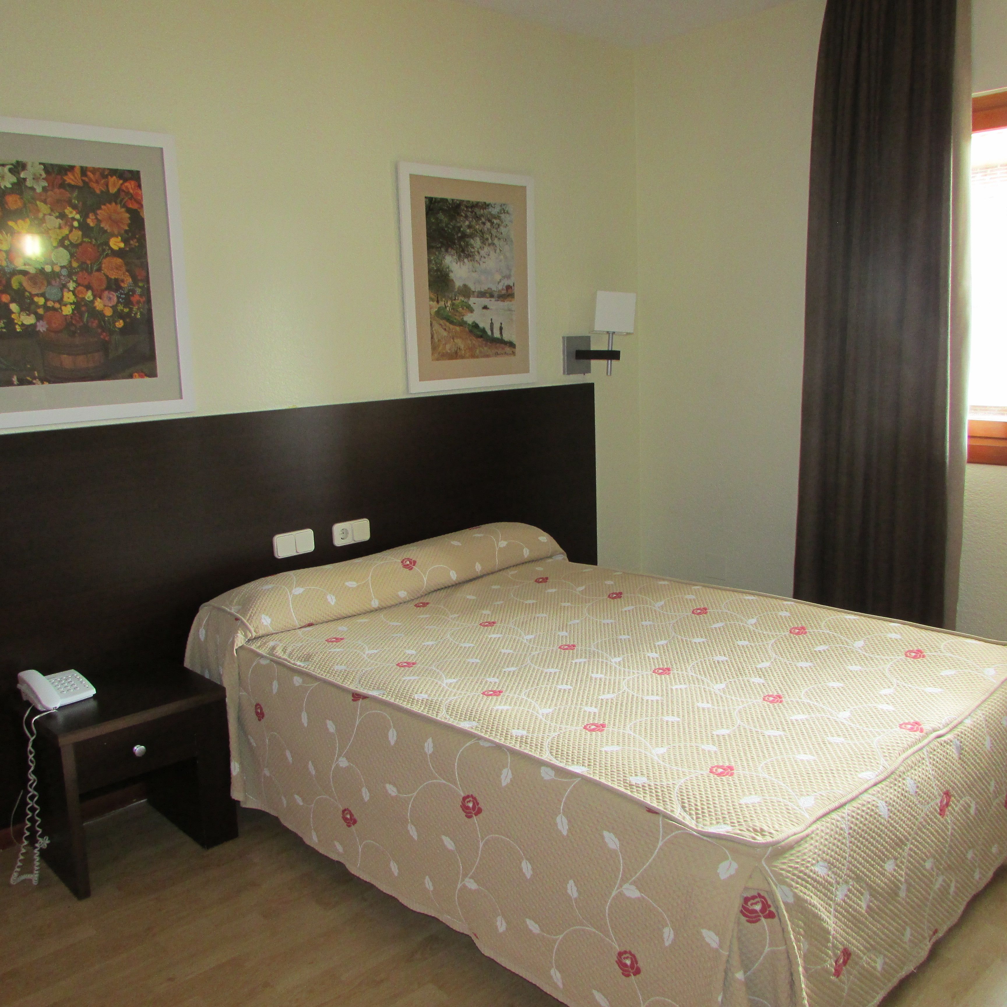 Habitación de hotel con una cama, una mesita de noche con teléfono, dos lámparas de pared y dos cuadros en la pared.