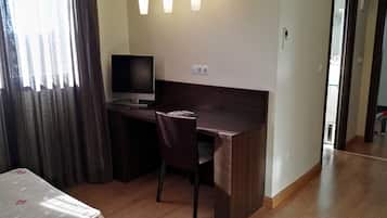 A small, well-lit room with a wooden desk, a chair, a television, and a bed with a floral bedspread.