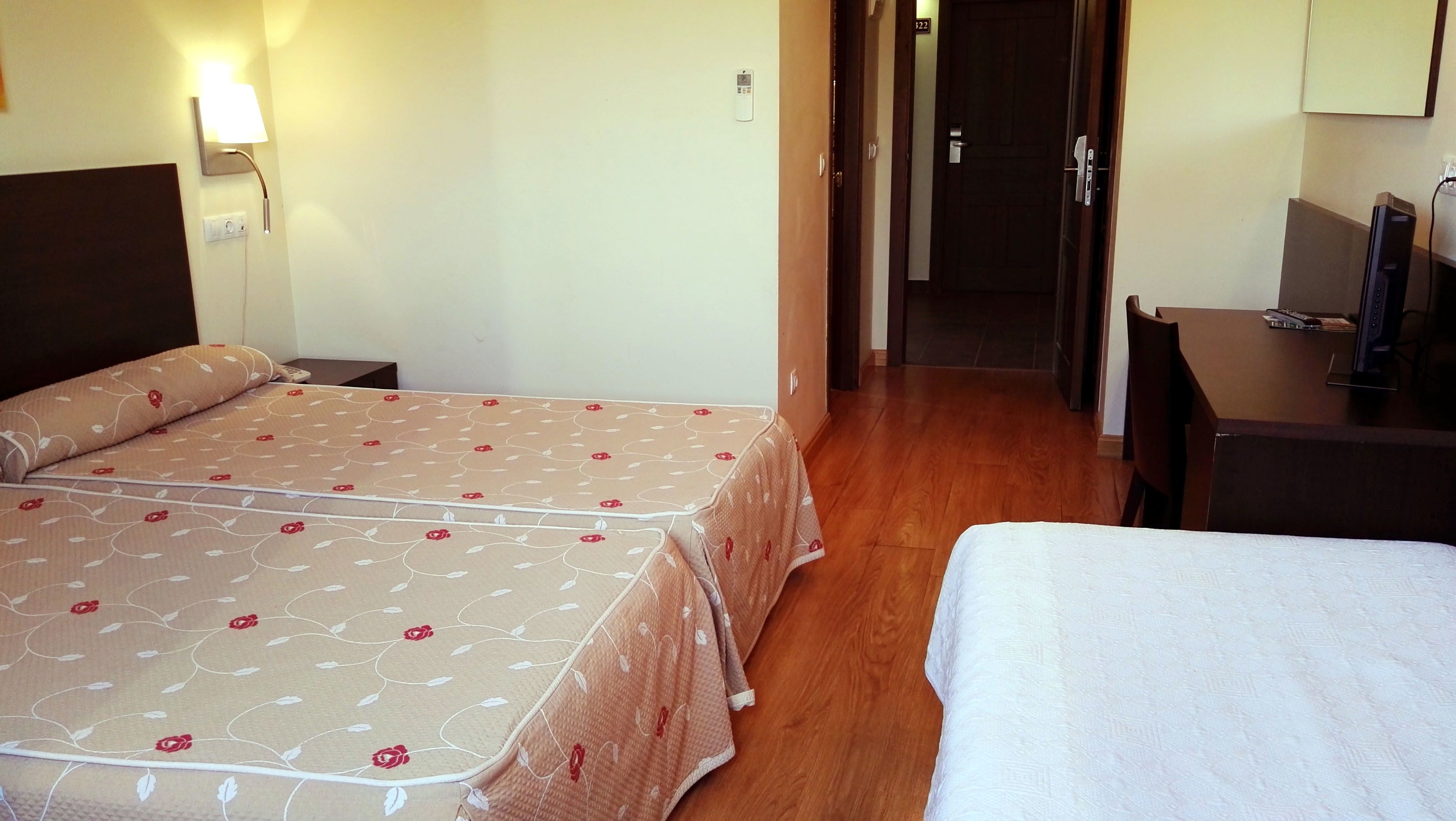 A hotel room with two beds, a desk, a TV, and a door leading to a hallway.