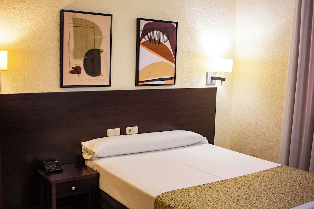 A hotel room with a bed, a nightstand, a phone, two framed artworks, and a wall-mounted lamp.