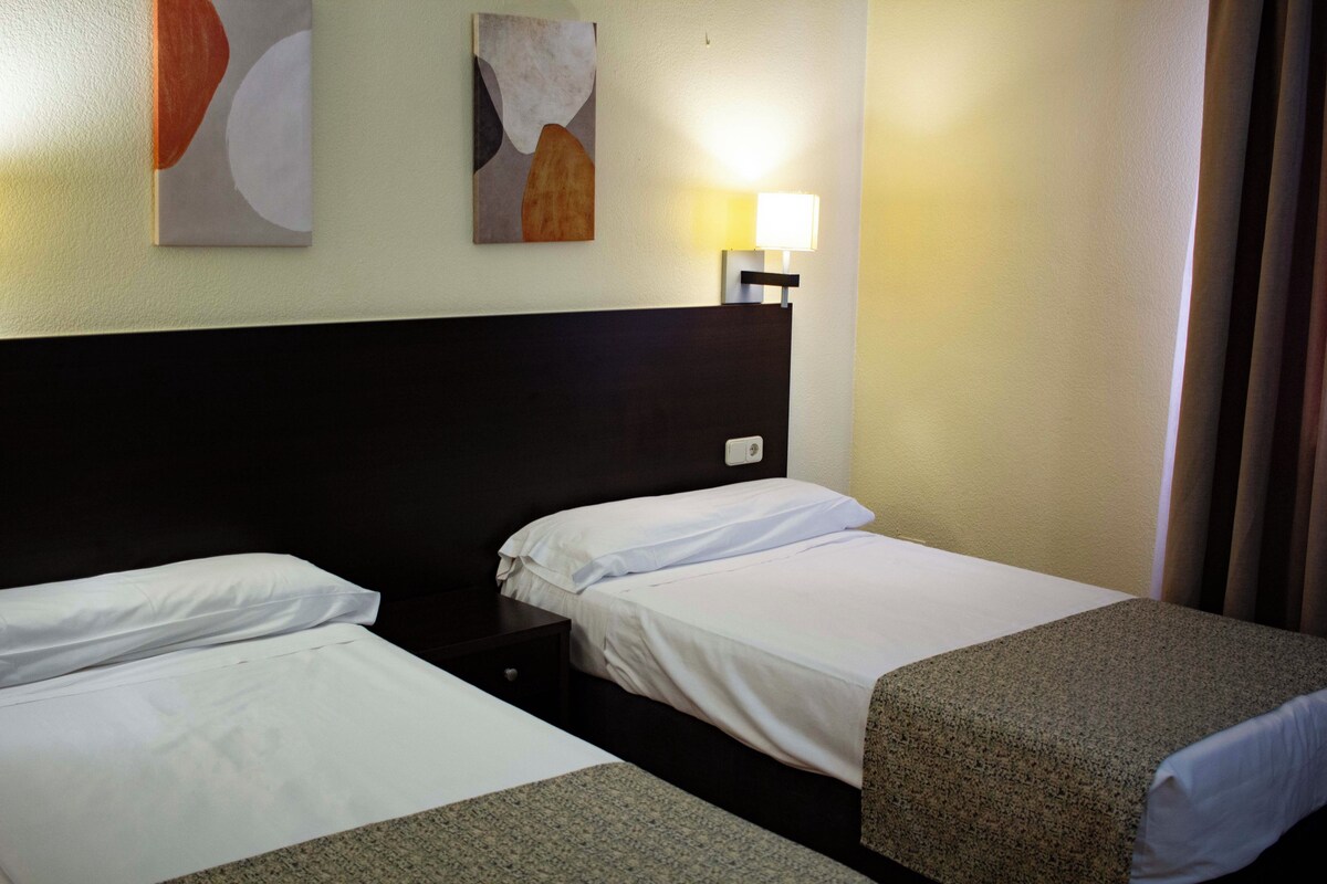 A hotel room with two single beds, a nightstand, a wall-mounted lamp, and abstract art on the wall.