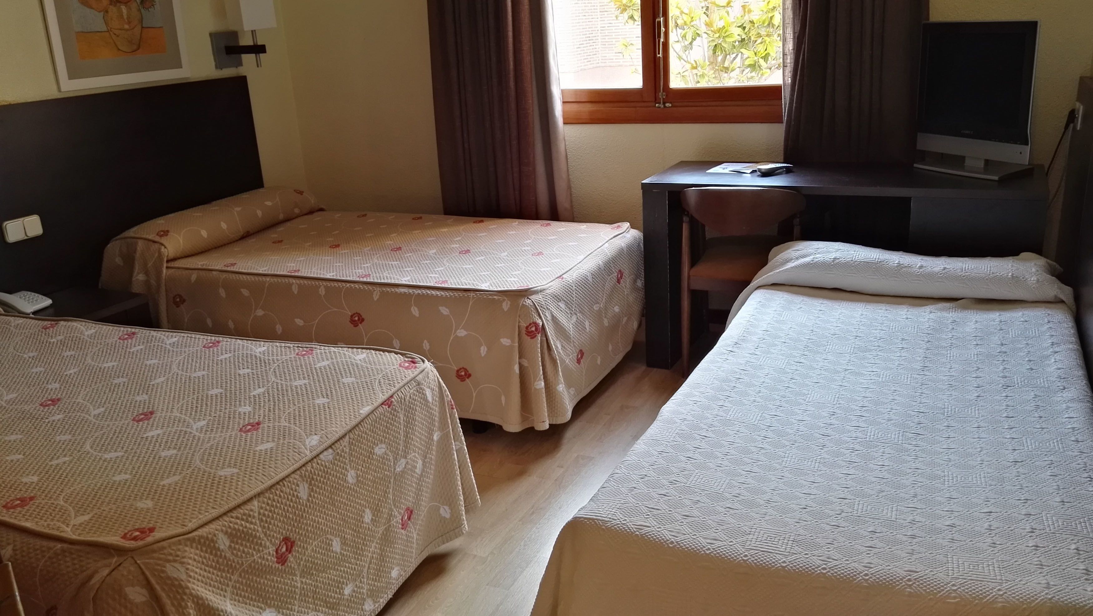 A hotel room with two single beds, a desk with a chair, a television, and a window with curtains.