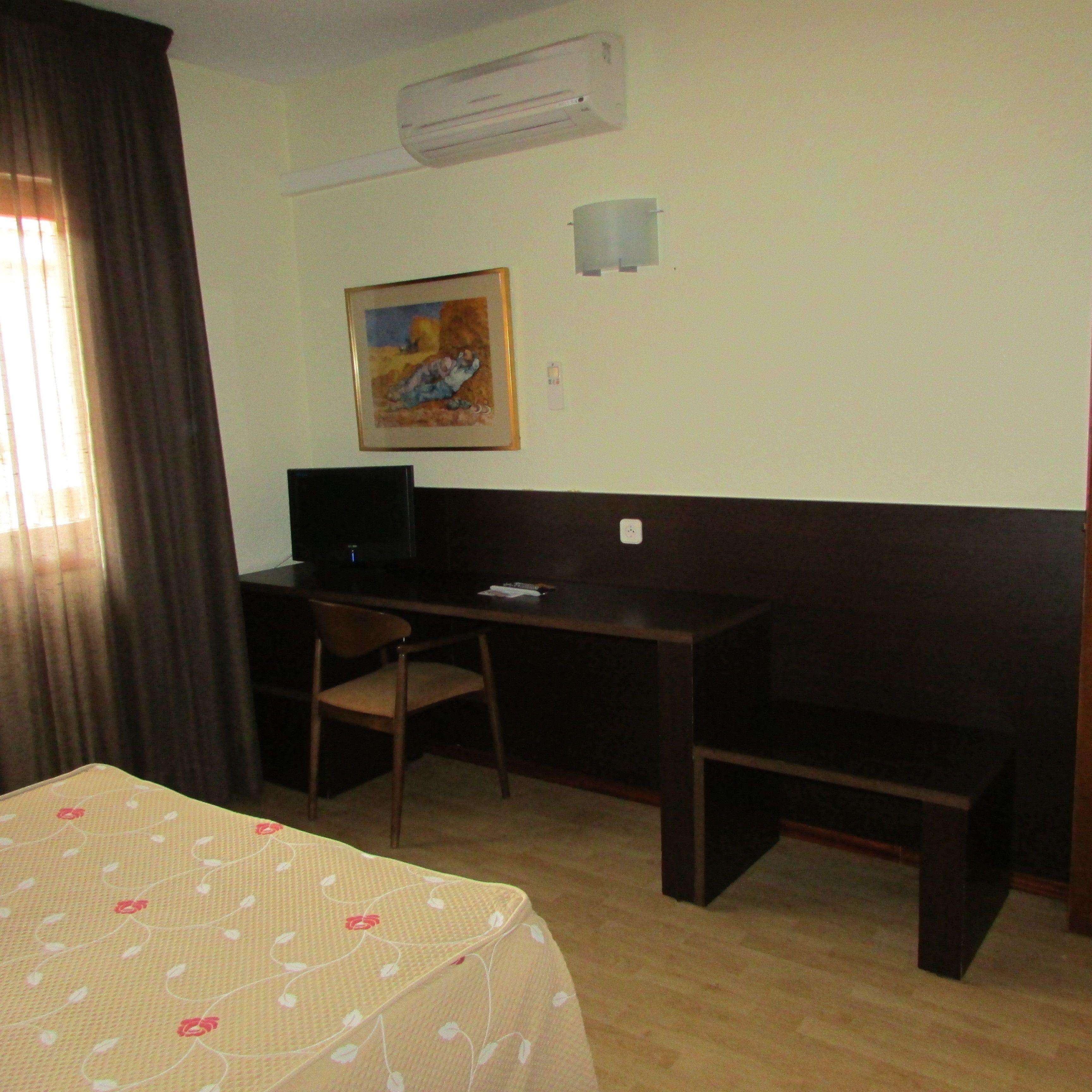 A hotel room with a bed, a desk, a chair, a bench, an air conditioner, and a painting on the wall.