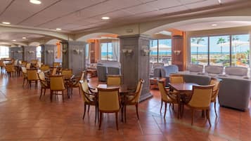 A spacious hotel lobby with multiple dining tables and chairs, a seating area with sofas, and large windows offering a view of the beach.