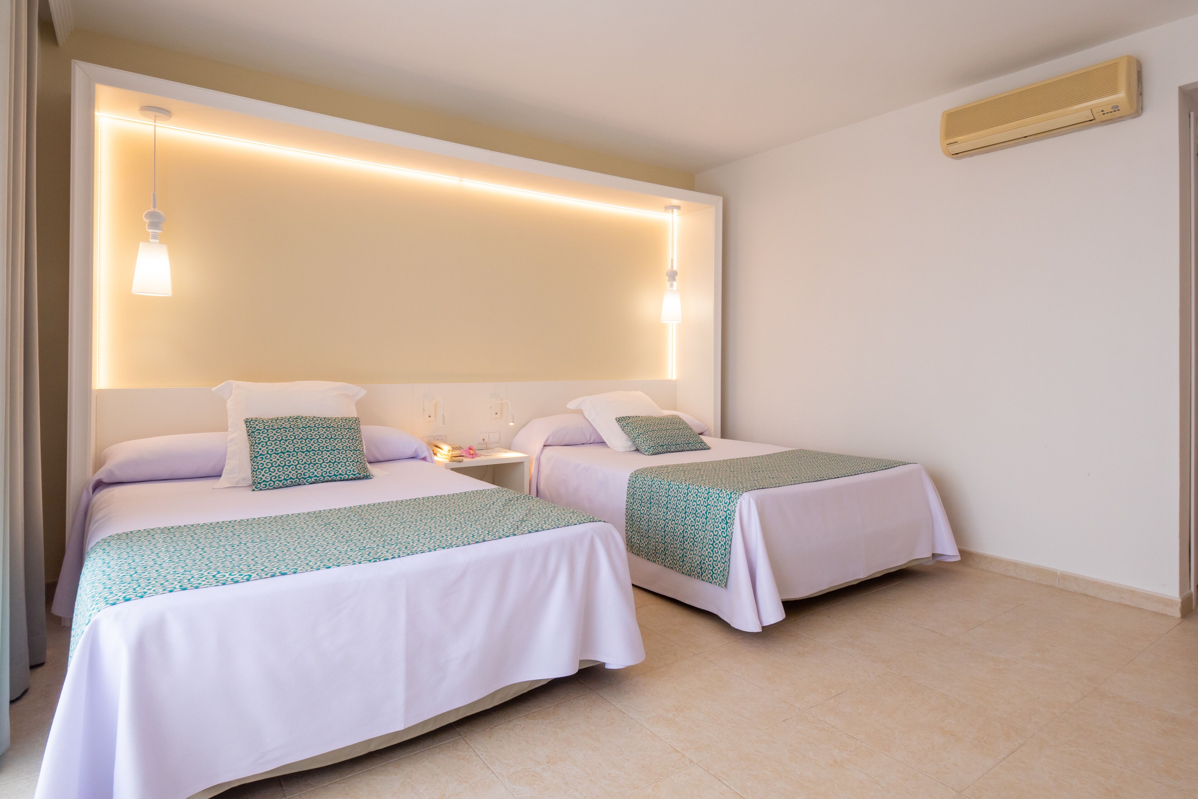 A hotel room with two single beds, each with a white bedspread and a green patterned pillow. There is a bedside table with a lamp and a small table lamp. The room has a modern design with a large window and a wall-mounted air conditioner.