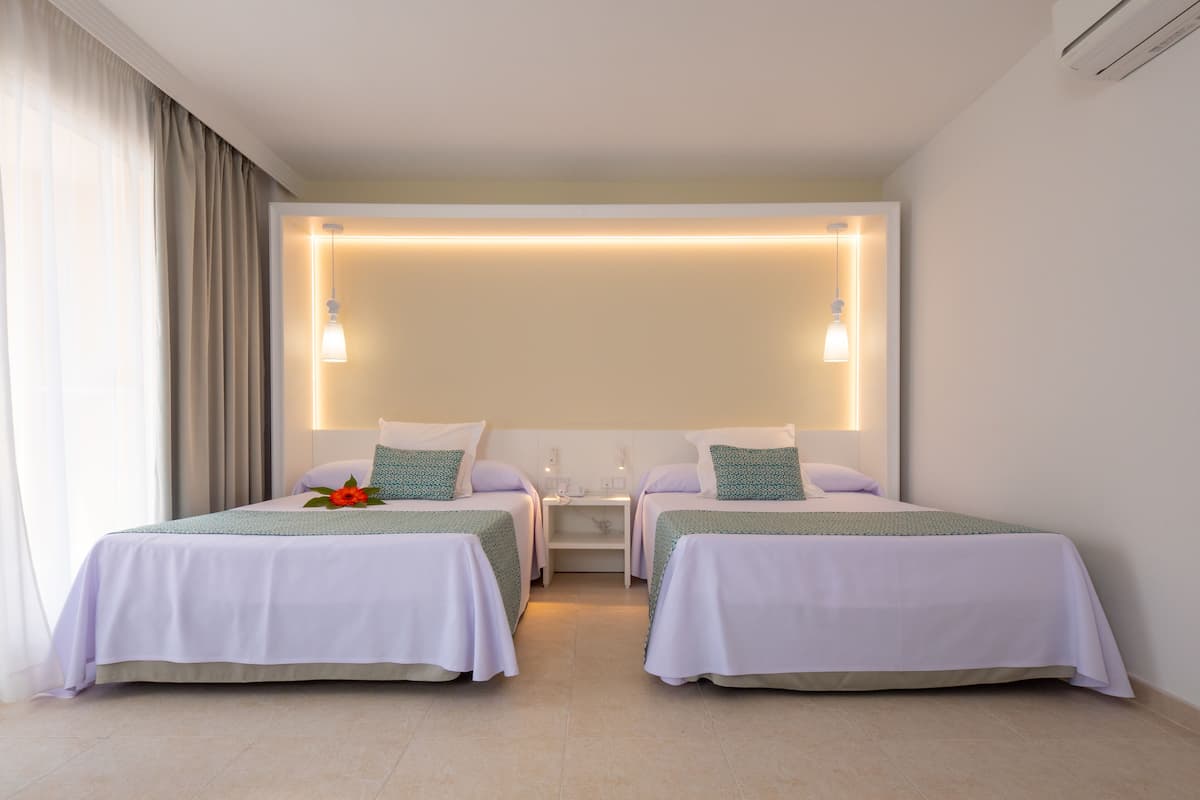 Une chambre d’hôtel avec deux lits simples, une table de chevet, un luminaire fixé au mur et une fenêtre avec des rideaux.