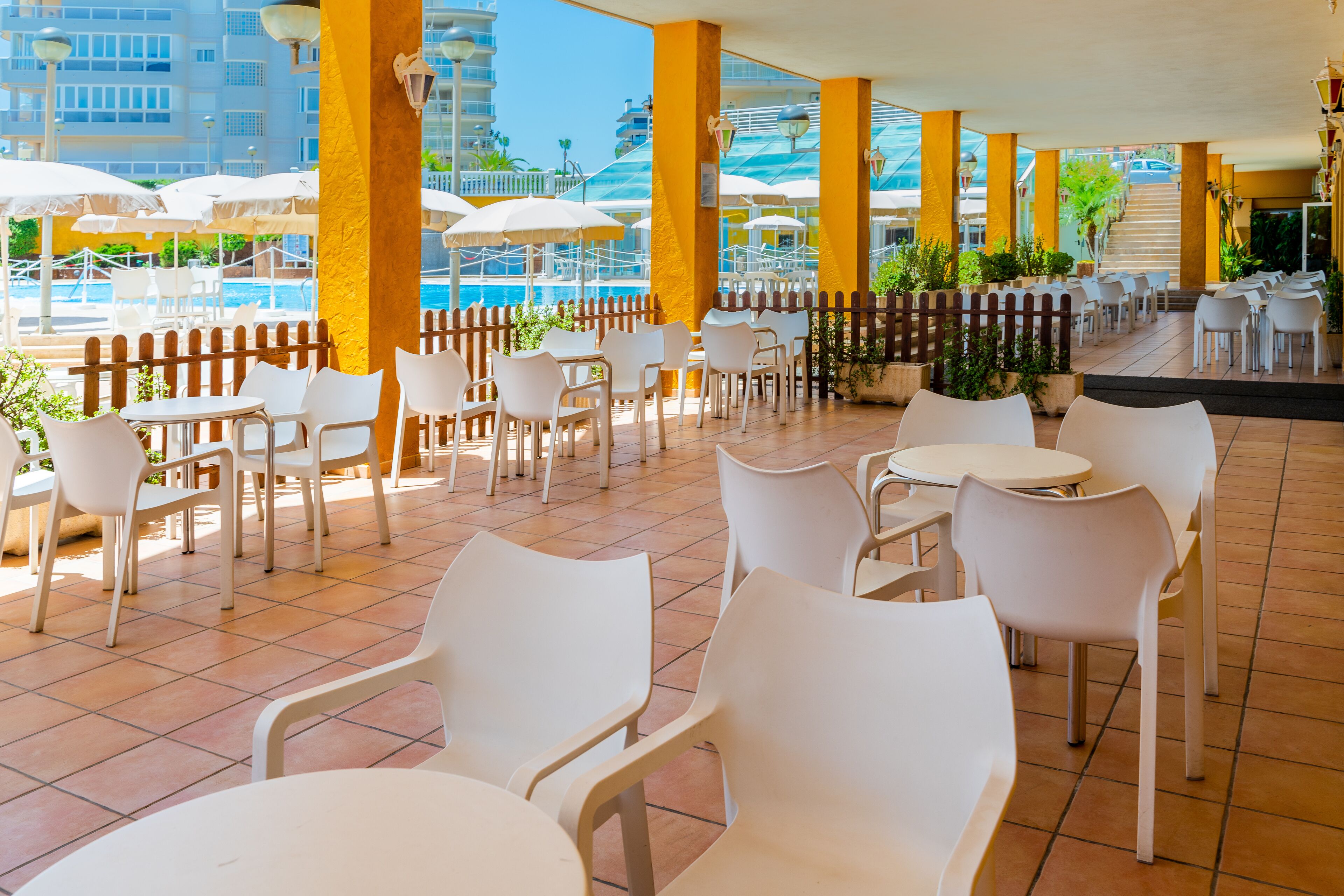 An outdoor patio with white plastic chairs and tables, yellow pillars, and umbrellas.