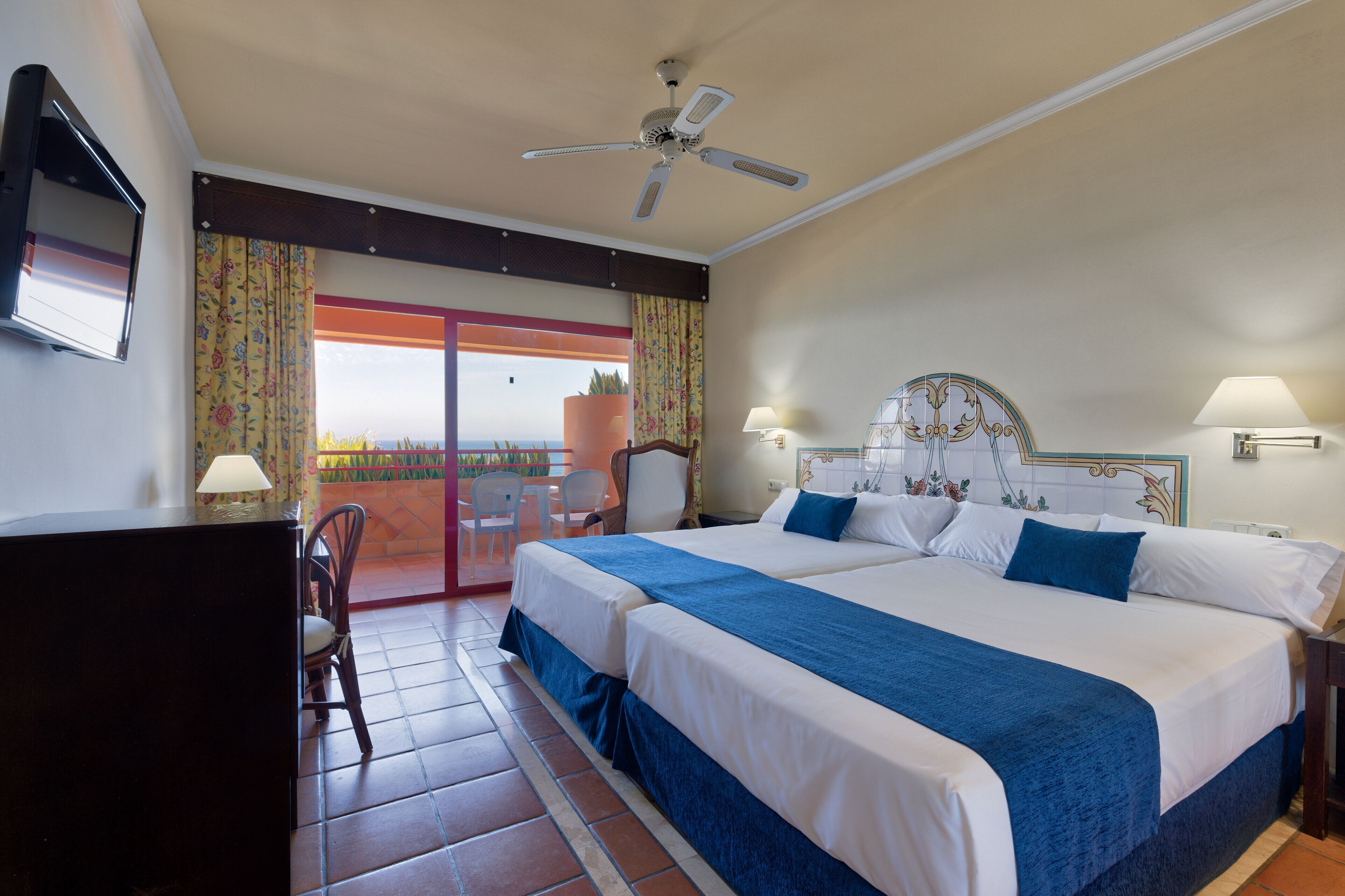 A hotel room with a large bed, a desk, a chair, a ceiling fan, and a view of the ocean.