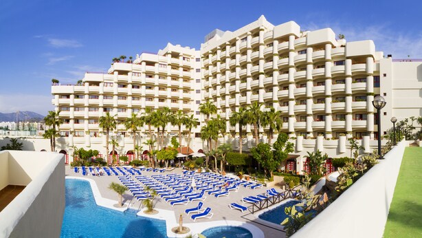 Un hotel con varios balcones, piscina con tumbonas y palmeras.